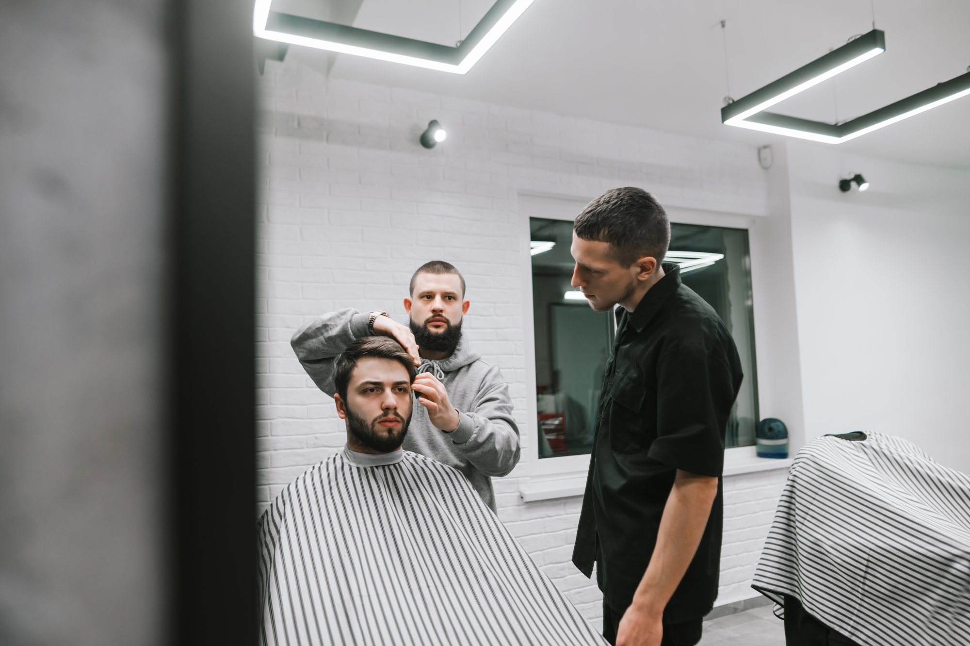 Two barbers create hairstyle for bearded man in bright modern hairdressing salon. Male model cut 2 hairdressers. Workshop on male haircuts in barbershop, training barber haircut